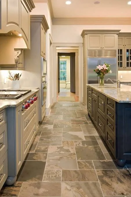 A modern kitchen with beige and dark blue cabinetry, granite countertops, stainless steel appliances, and a flower vase on the island. There's a doorway at the end leading to another room with a window showing greenery outside.