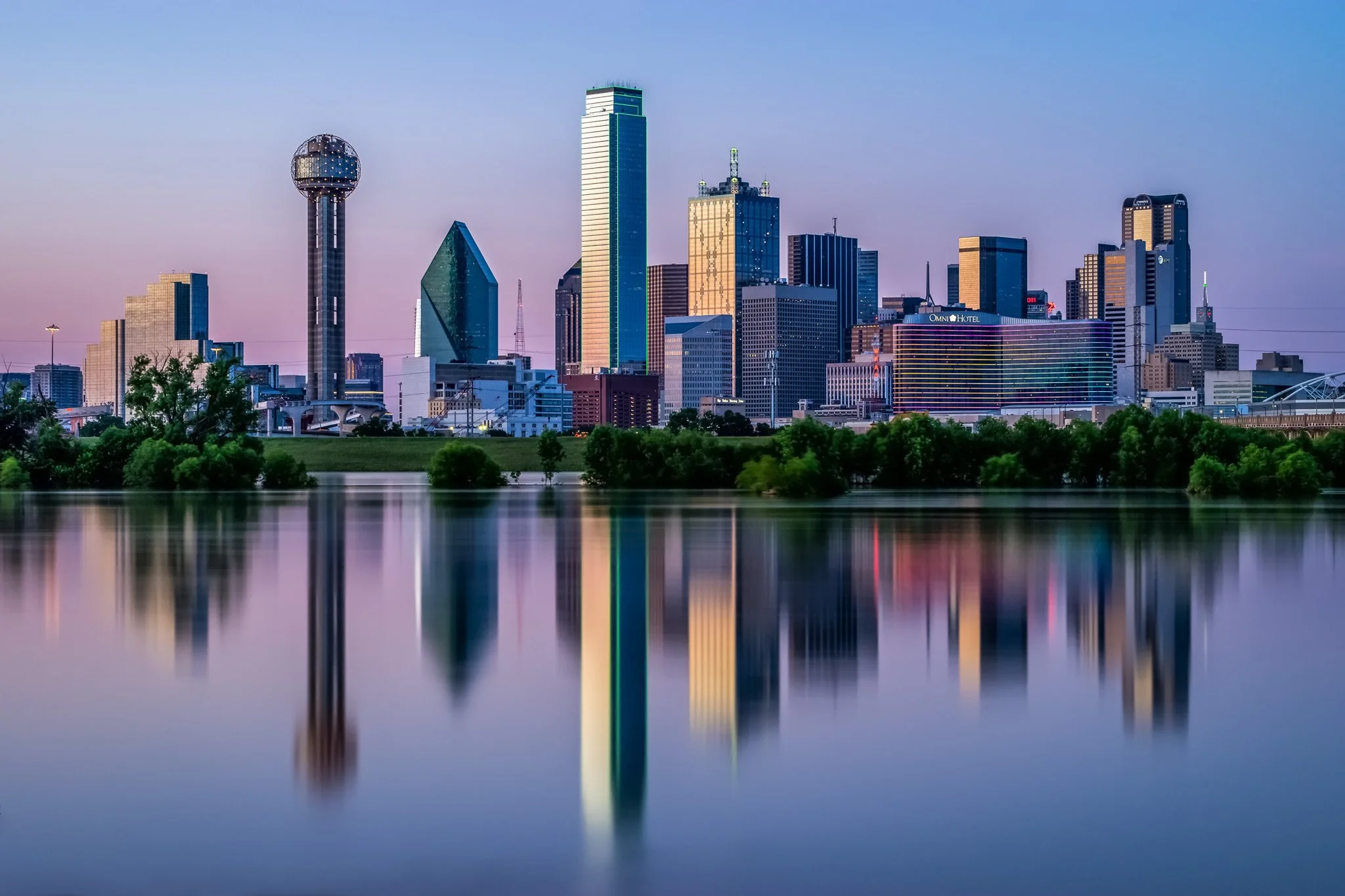 Dallas skyline at sunset with reflections in the water, Central Expressway Tower, and Omni Hotel visible.