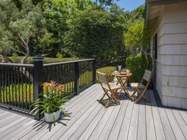 A wooden outdoor deck with a black railing, a small round table with two chairs, and a potted plant, surrounded by lush green trees and bushes visible in the background, in Frisco, Texas.