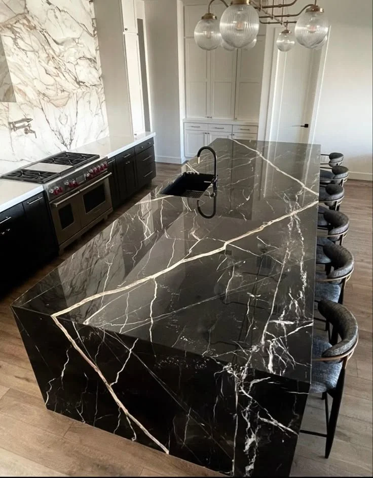 A black marble kitchen island with white veining, situated in a modern kitchen with dark cabinets, a stove, a sink, a white marble wall, and pendant lights overhead.