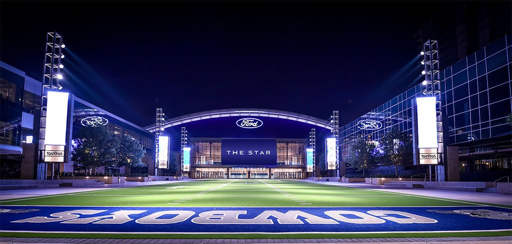 STAR Ford stadium at night with illuminated signs, a large LED screen displaying "The Star," a football field, and surrounding glass buildings.