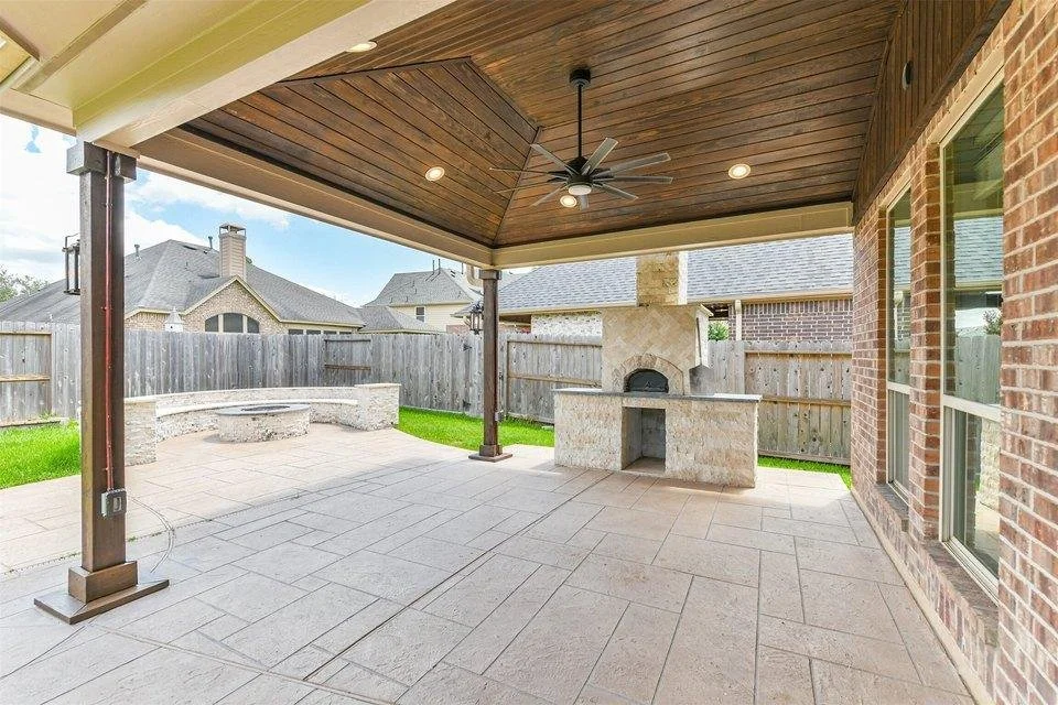 Covered patio with a ceiling fan, built-in brick oven, and outdoor fireplace, enclosed by a wooden fence, with neighboring houses visible in the background.