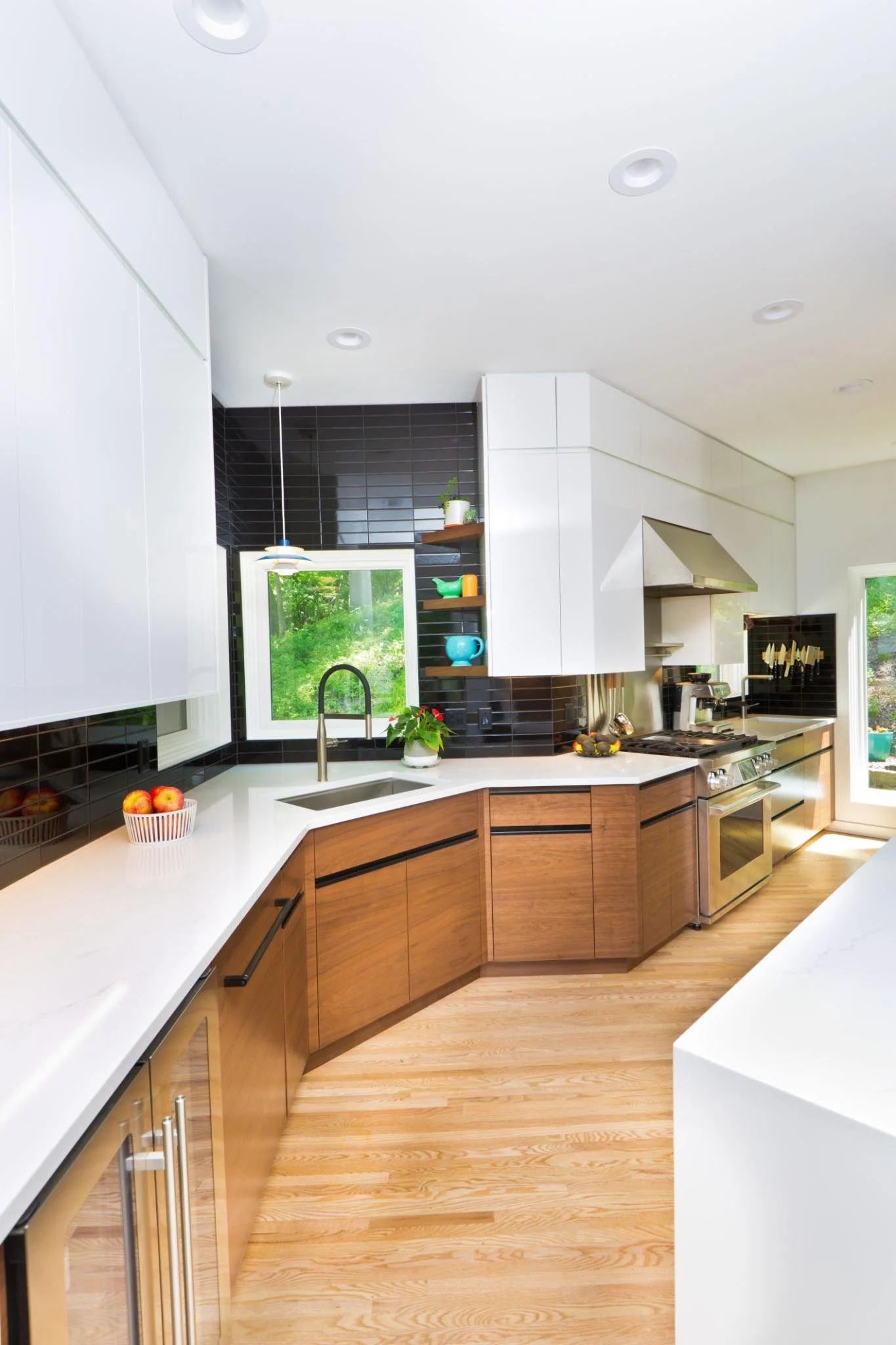 Modern kitchen with white countertops, wooden cabinets, black tile backsplash, stainless steel appliances, and large windows showing greenery outside.