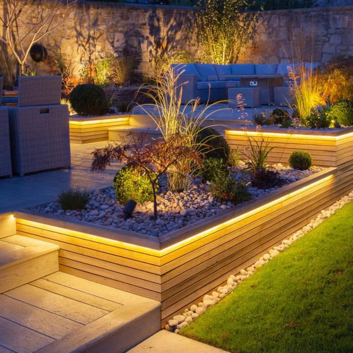 Nighttime view of a modern backyard garden remodel done in Prosper, Texas with illuminated LED lighting along wooden raised garden beds filled with plants and rocks, with outdoor furniture in the background.