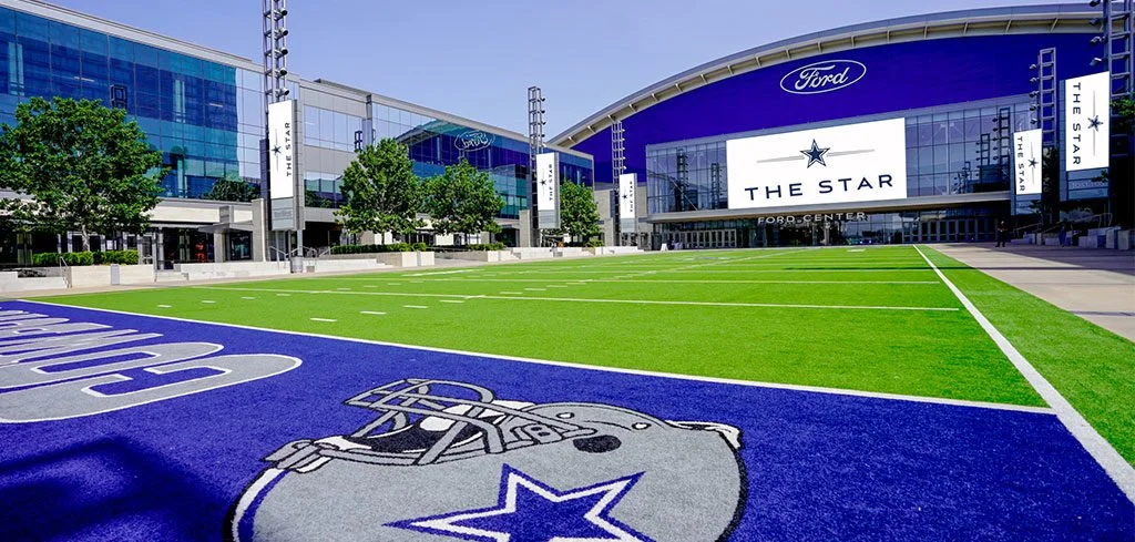 Exterior of the Ford Center at The Star football training facility with a football field and Dallas Cowboys logo, surrounded by glass buildings and trees.