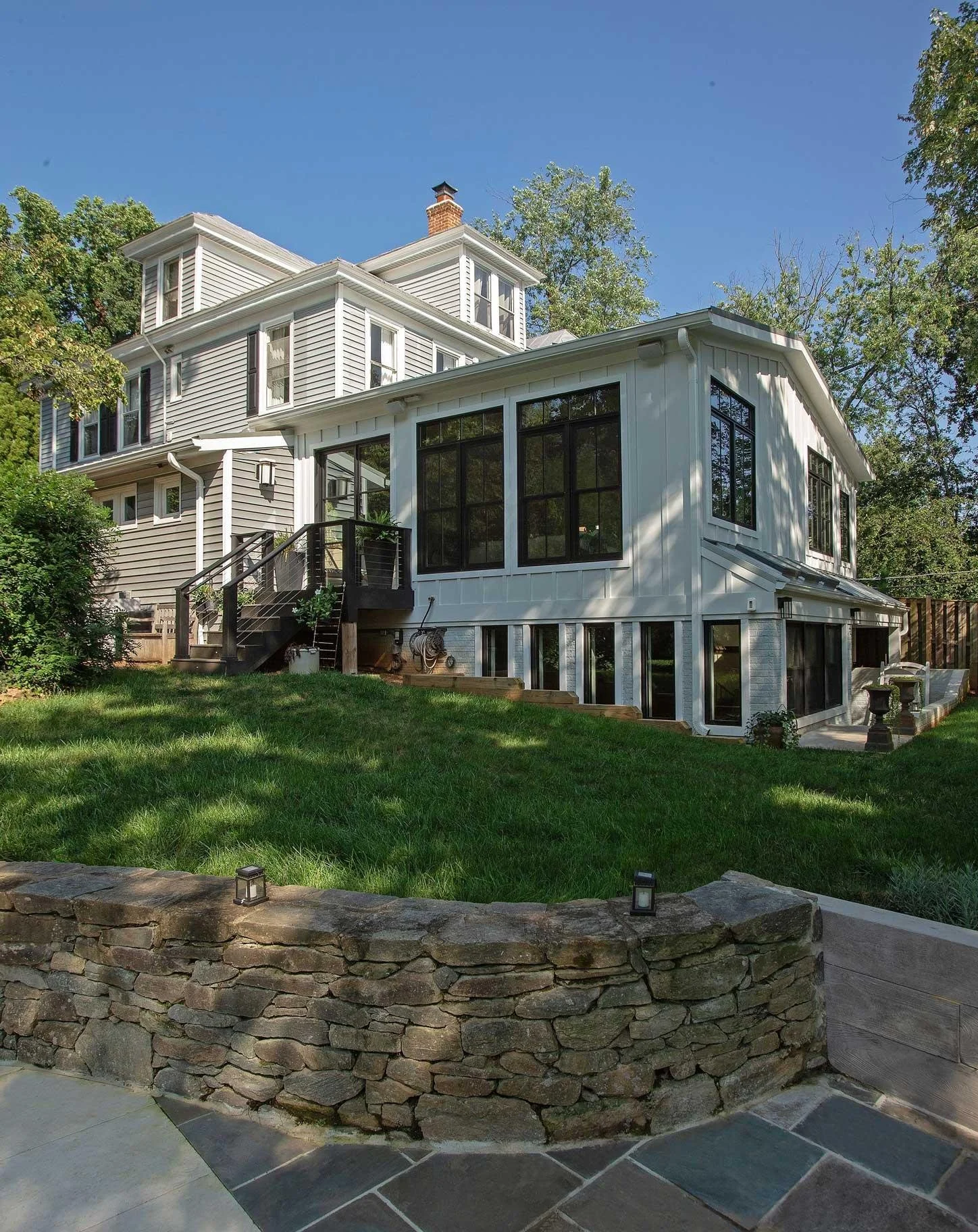 A multi-story house with white siding and large black-framed windows, a wooden staircase leading from the backyard to the house, a stone retaining wall in the foreground, green grass, trees surrounding the house, and a clear blue sky.