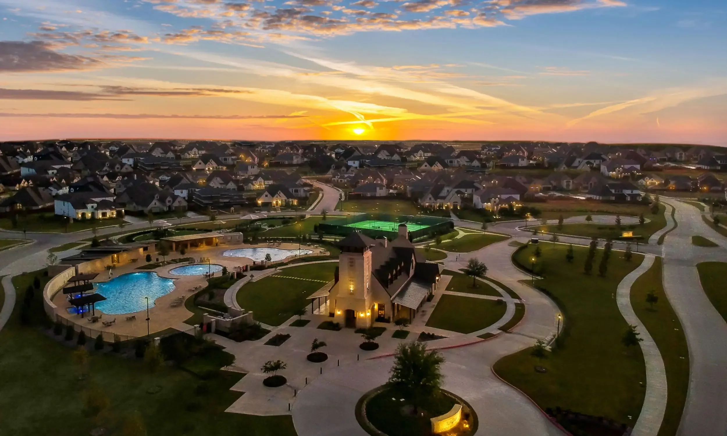 A neighborhood at sunset with houses, a community pool, tennis courts, and paved walkways.