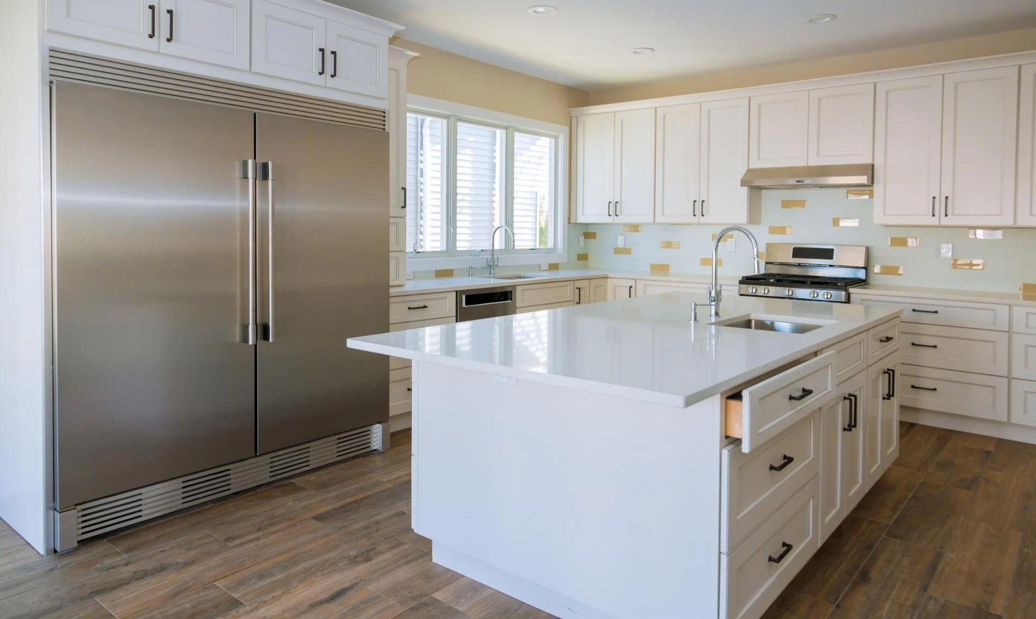 A modern kitchen with white cabinets, a white island countertop, stainless steel appliances, and wooden flooring. There is a large window above the sink with sunlight coming in.