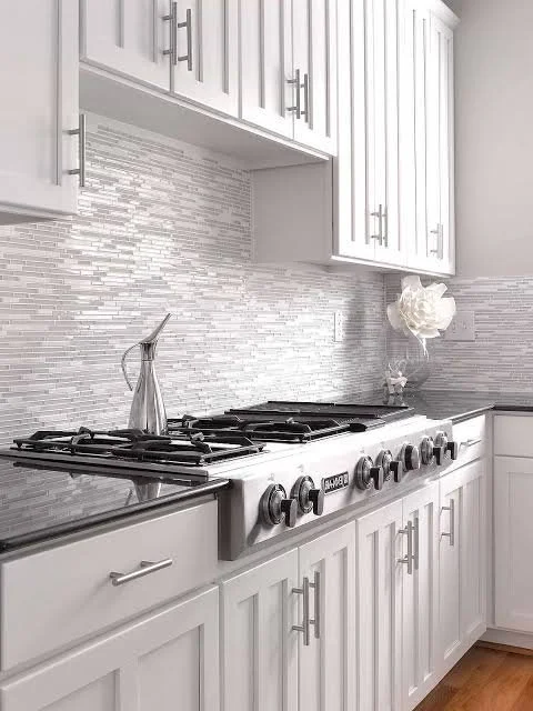 Modern white kitchen with a stainless steel gas stove, backsplash with horizontal tile pattern, and upper and lower cabinets with silver handles. There is a silver pitcher and a decorative white flower in a clear vase on the countertop.