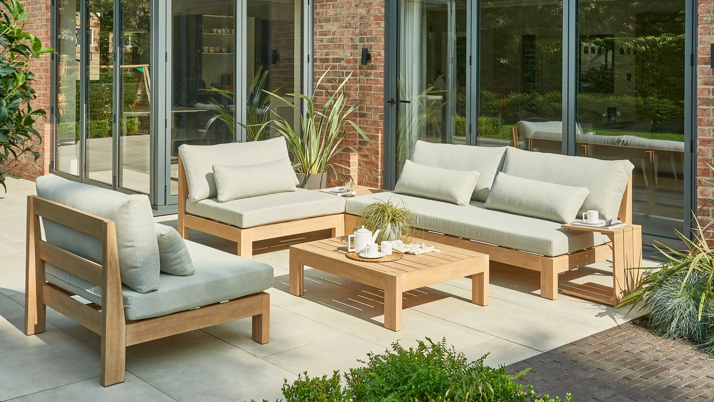 An outdoor patio remodeling project with modern beige cushioned furniture, including two sofas and a armchair, arranged around a wooden coffee table with plants and cups. There are large glass sliding doors and potted plants.