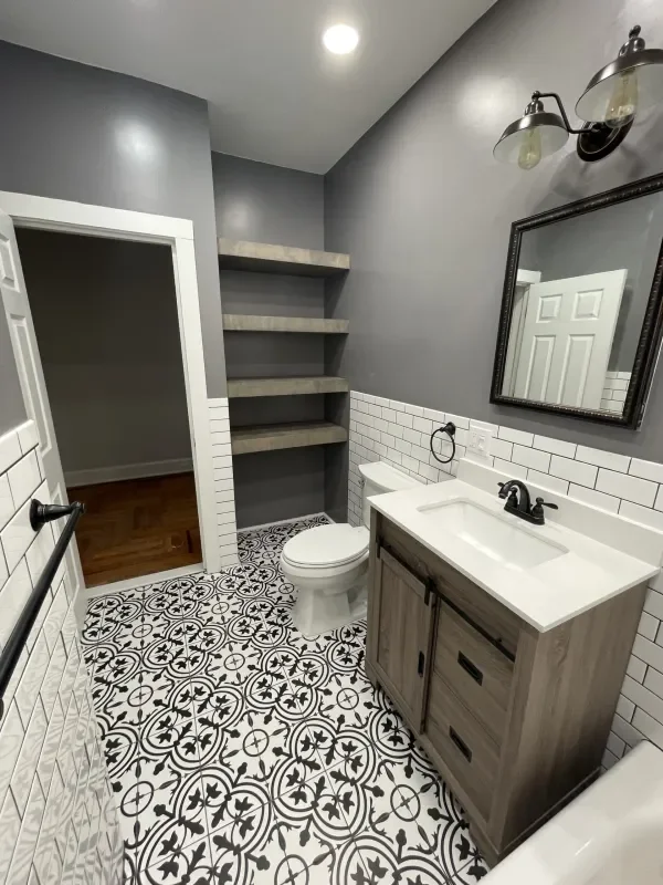 A small bathroom with gray walls, patterned black and white tile flooring, a white toilet, a wooden vanity with a white sink, a black faucet, a mirror with a black frame, a sconce light fixture, white subway tiles on the lower half of the wall, and three floating gray shelves on the wall.