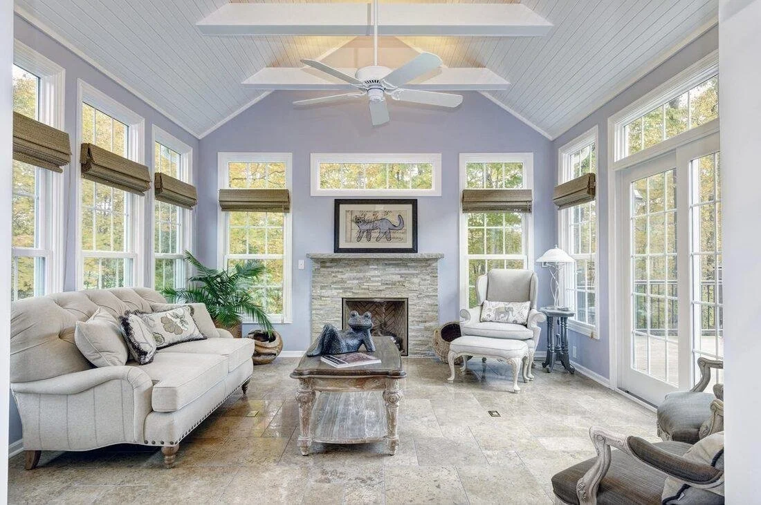 Bright living room with large windows, a stone fireplace, and light-colored furniture including a sofa, armchair, and side table with a lamp.