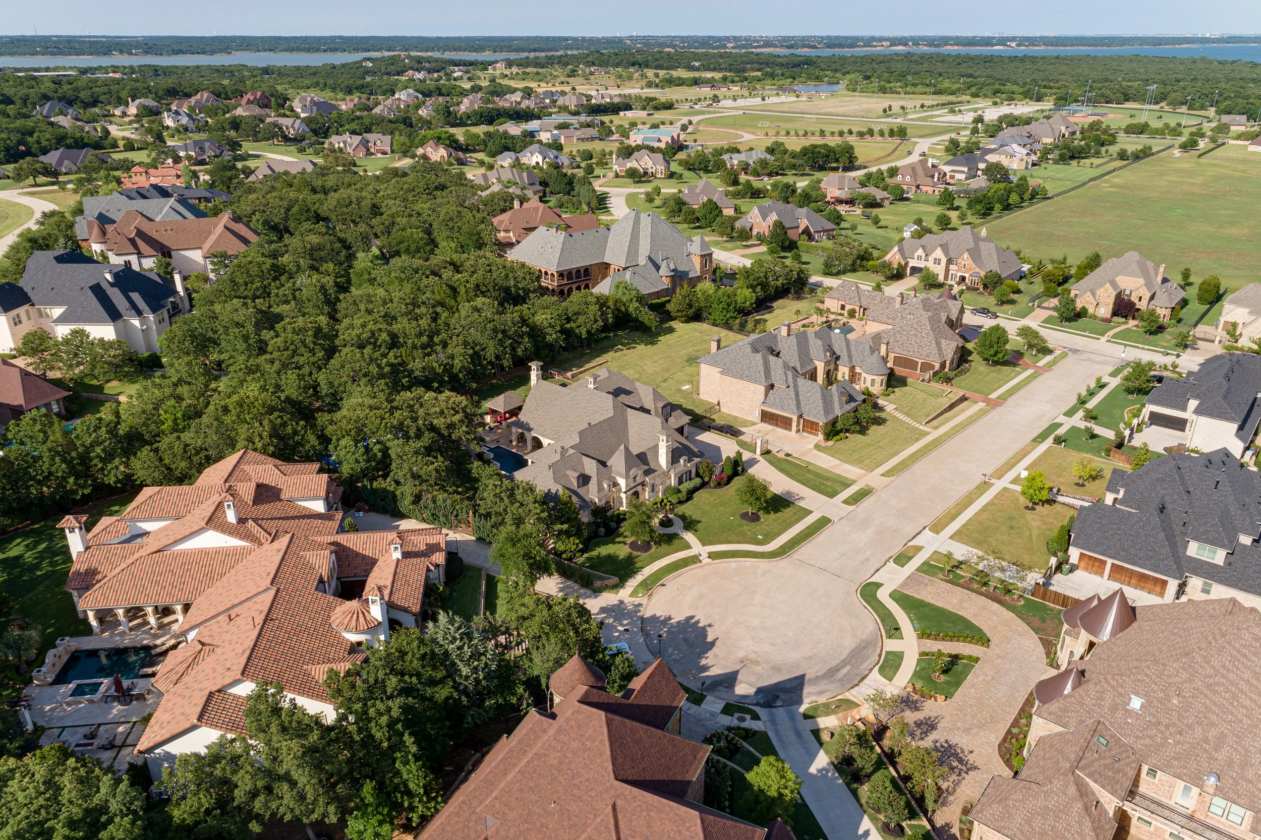 Aerial view of a suburban neighborhood with large houses, trees, and a mix of paved and grassy areas. In the background, a lake and sports fields are visible.