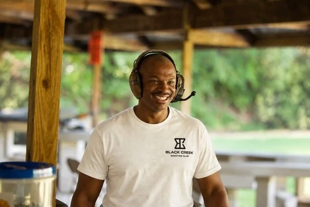 A smiling man wearing a white T-shirt that says "Black Creek Shooting Club" and a headset with a microphone, outdoors under a wooden shelter with greenery in the background.
