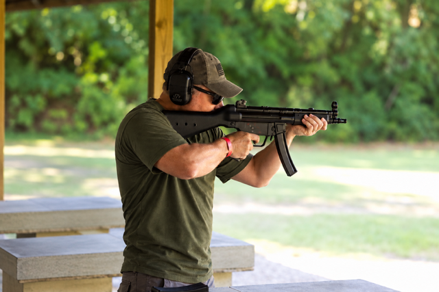A man at a shooting range aiming a rifle, wearing ear protection and a cap.