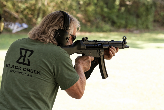 Man with long gray hair wearing headphones, aiming a rifle outdoors.