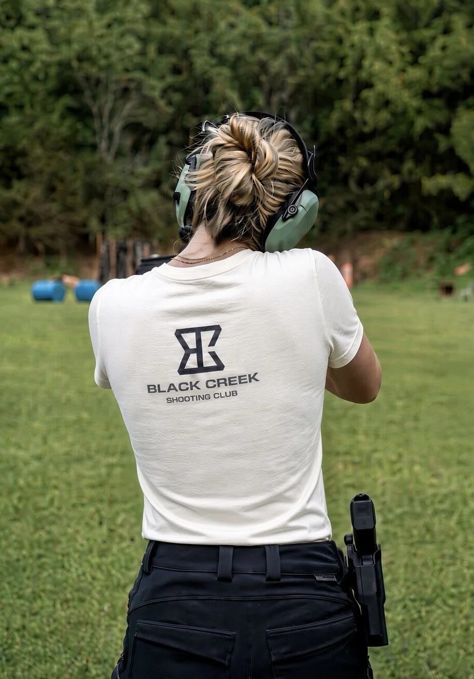 A woman at a shooting range wearing a White T-shirt with 'Black Creek Shooting Club' logo, black pants, and hearing protection, aiming with a handgun.