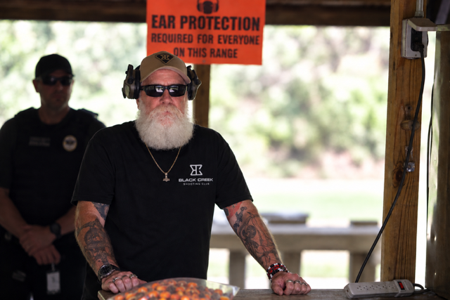 A bearded man wearing sunglasses, headphones, and a black t-shirt with the logo 'Black Creek Shooting Club' stands at a table with his hands resting on it. Behind him is a red sign that says 'Ear Protection Required for Everyone on This Range.' In the background, a police officer is observing, and the scene is outdoors with blurred greenery visible.