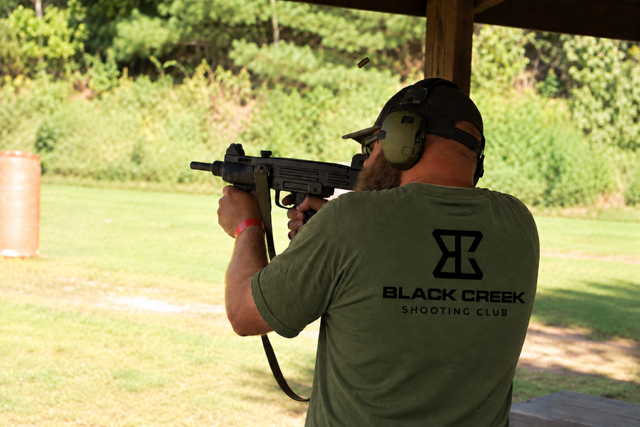 A man at a shooting range aiming a firearm, with headphones on and wearing a green T-shirt with 'Black Creek Shooting Club' printed on the back.