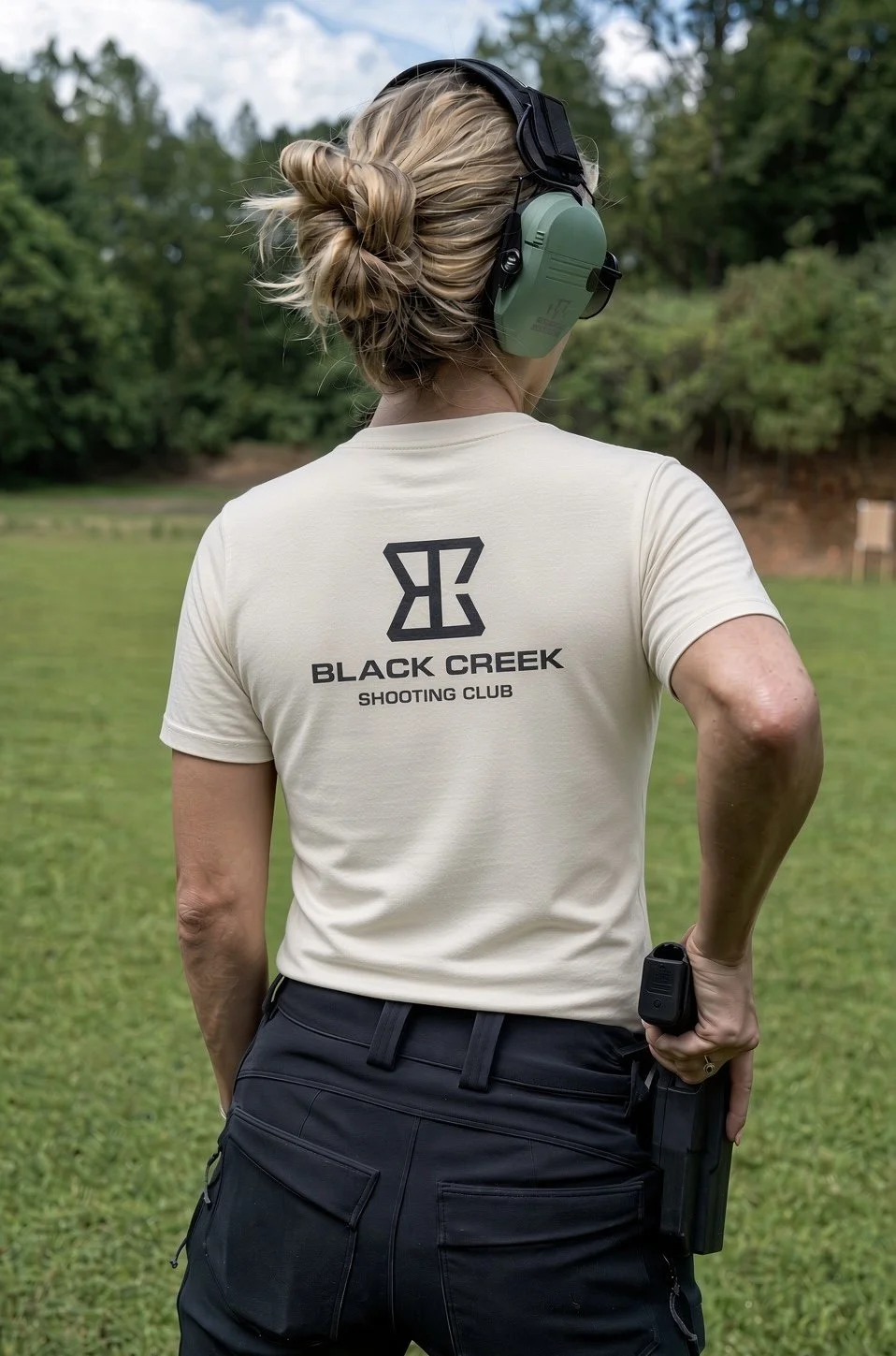 A woman with blond hair in a bun wearing a beige t-shirt with 'Black Creek Shooting Club' and a logo on the back, standing outdoors on a grassy field with trees in the background, holding a handgun behind her back.