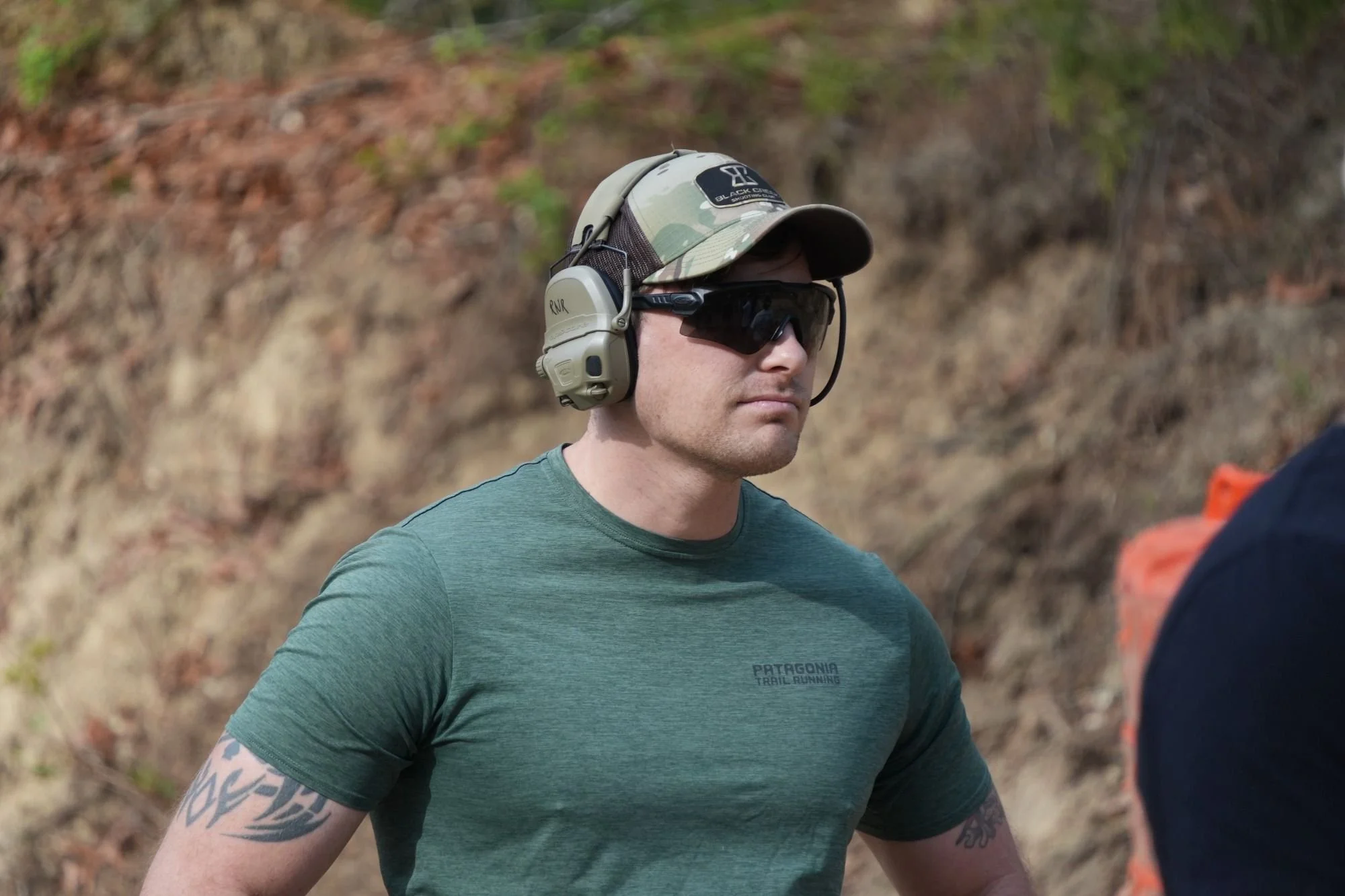 A man wearing tan tactical headphones, black sunglasses, a dad hat with camouflage patterns, and a teal t-shirt, standing outdoors in a natural setting with trees and soil in the background.