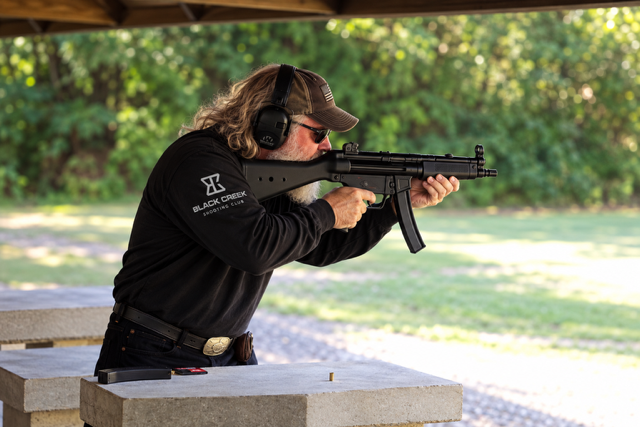 Man aiming a rifle at outdoor shooting range with trees in the background.