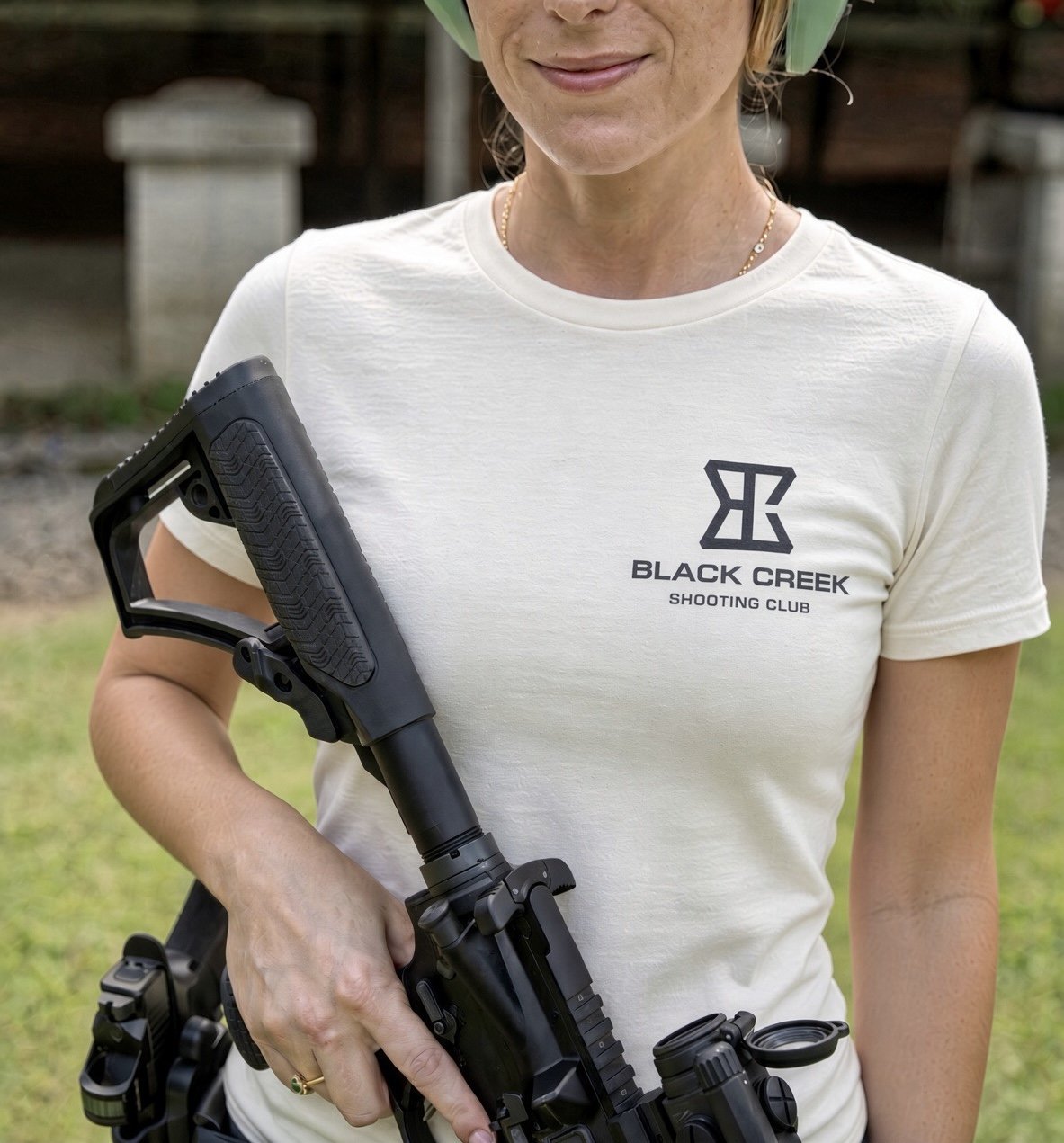A woman wearing a white t-shirt with 'Black Creek Shooting Club' logo, holding a firearm with a sight.