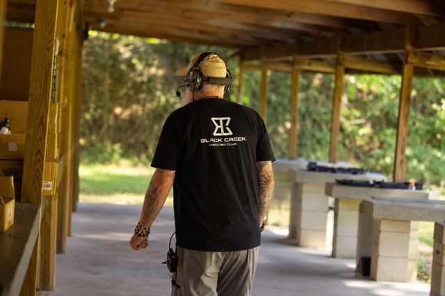 Man wearing a black t-shirt, beige cap, and headphones walking at an outdoor shooting range with rifles on tables in the background.