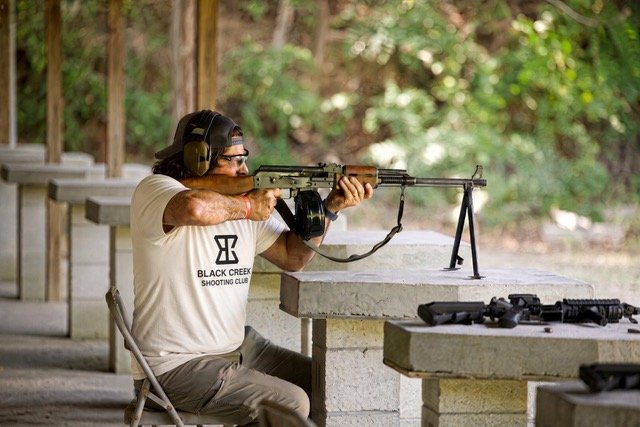 Man at outdoor shooting range aiming rifle while wearing hearing protection and glasses, seated at concrete shooting table with guns nearby.