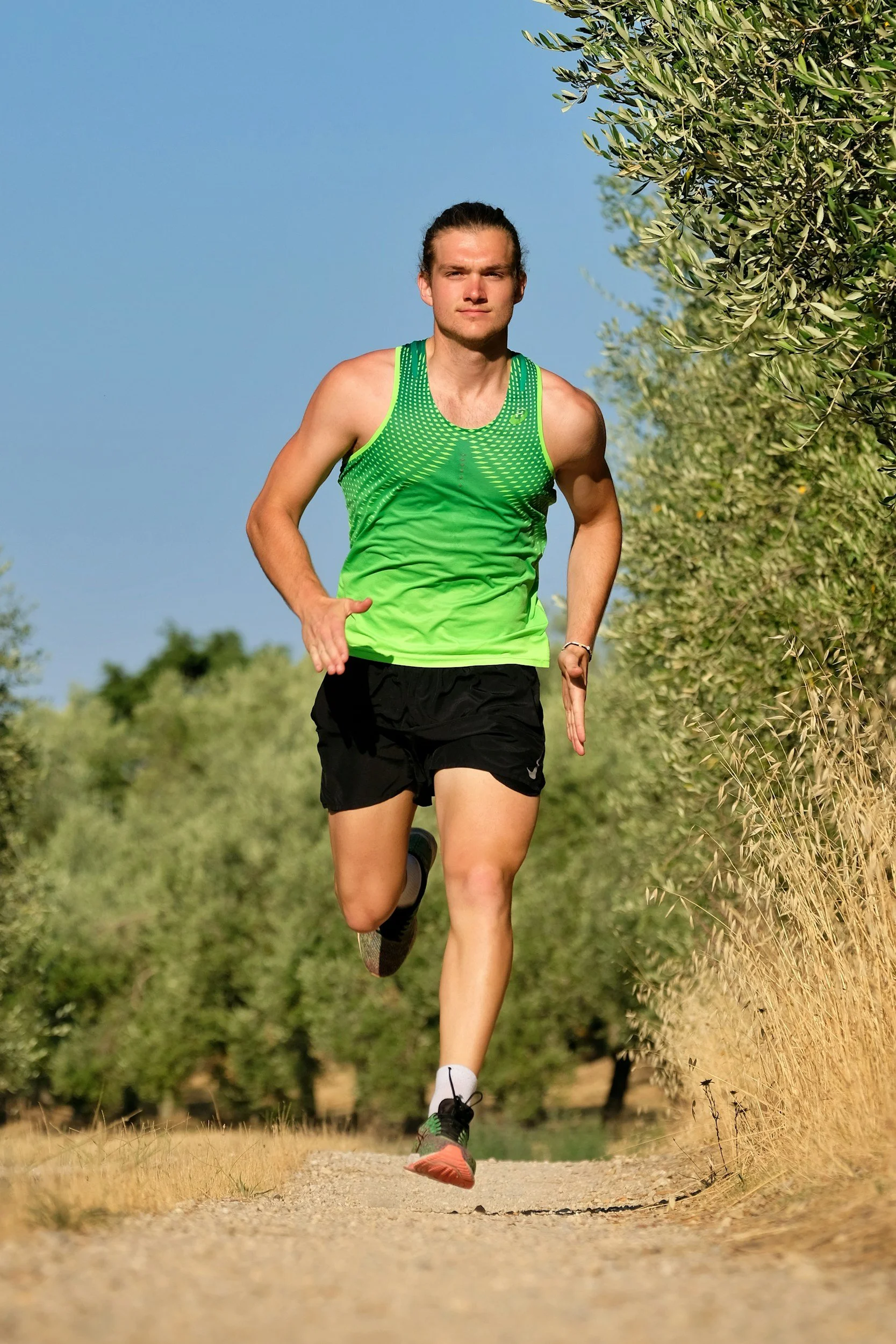 Young man running on dirt trail surrounded by greenery, wearing a bright green athletic tank top and black shorts, with a focused expression.