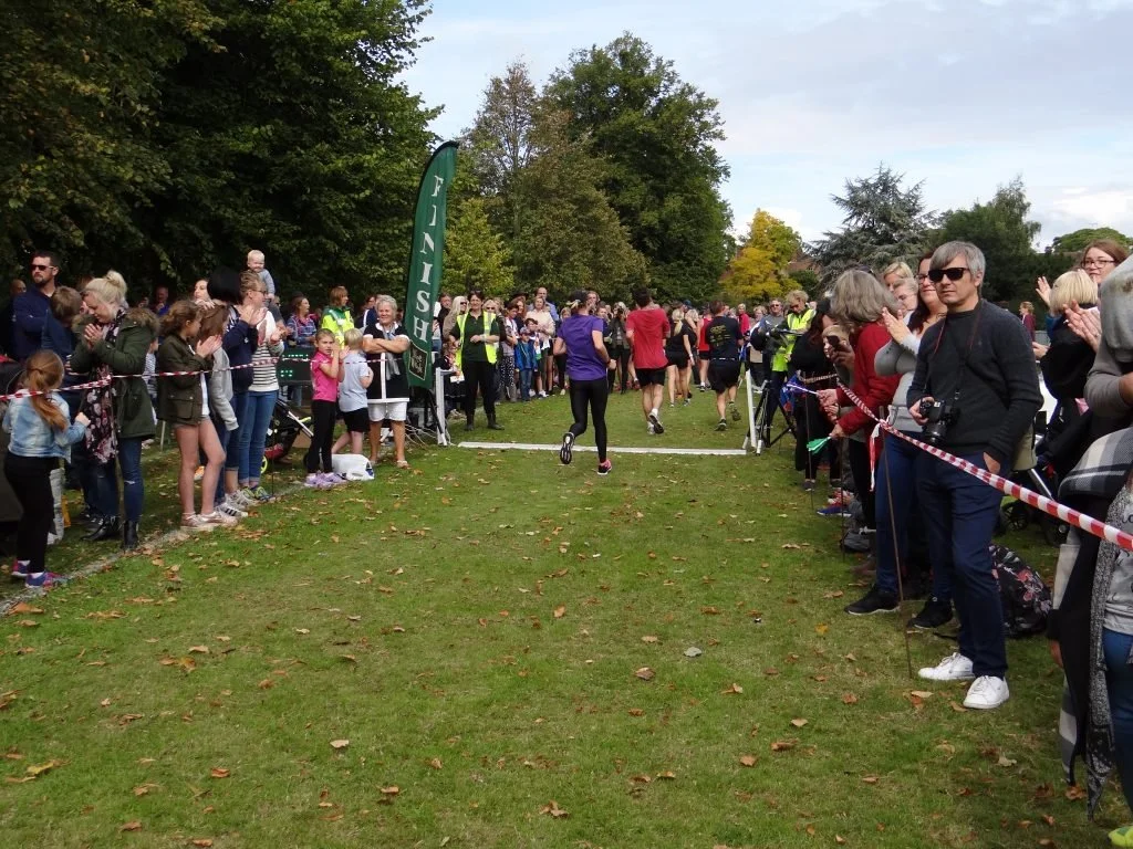 People at a park crossing the finish line of a race, with spectators on both sides clapping and cheering.