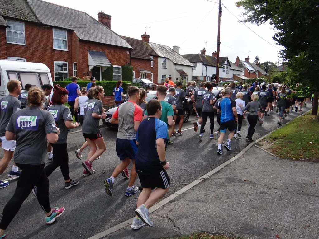 A large group of people participating in a community race or fun run on a residential street, with houses and trees in the background.