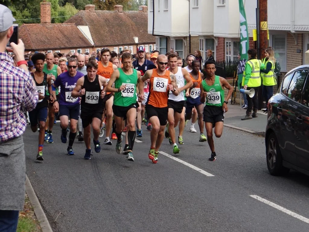 A group of runners participating in a road race, some wearing numbered bibs, running on a street lined with houses and spectators. A person in the foreground is taking a photo, and there are event volunteers in yellow vests on the sidewalk.