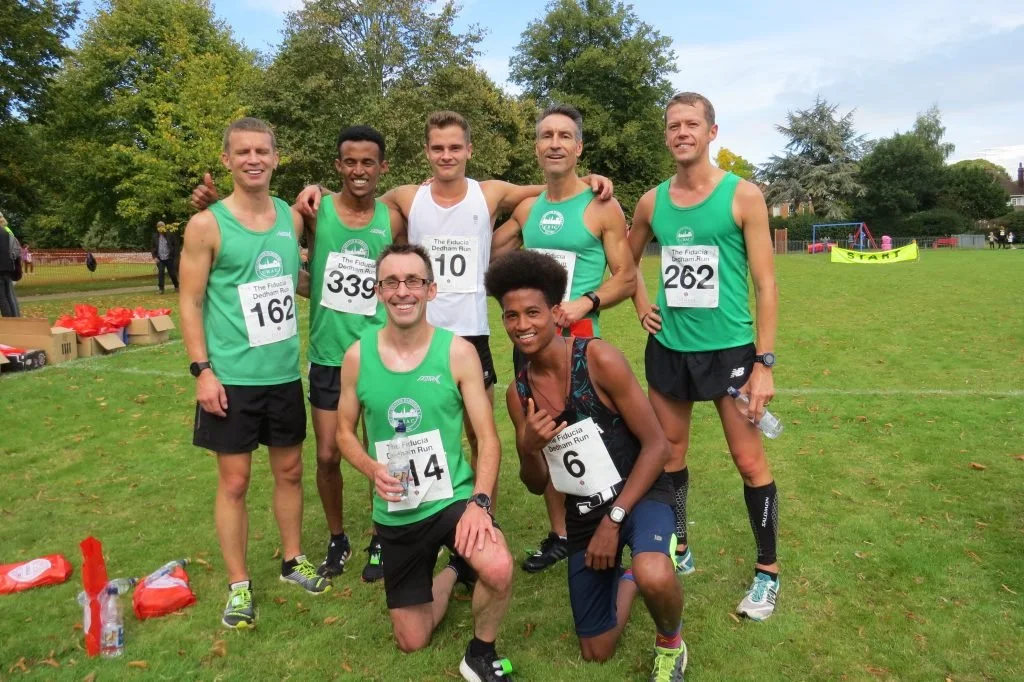 A group of seven runners posing together after a race in a park. They are wearing athletic gear with race bibs, and trees and a playground are visible in the background.