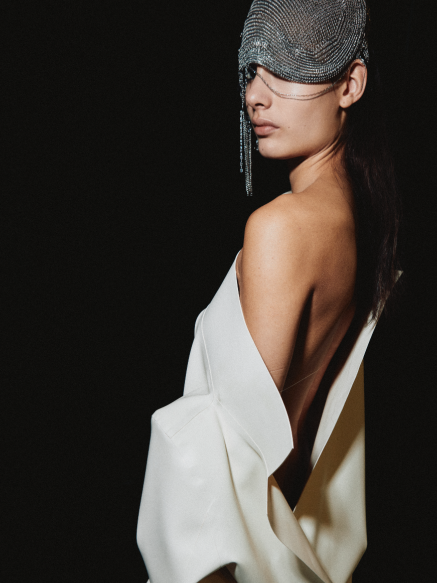 A woman with a silver beaded headpiece and face covering, dressed in a stylish off-the-shoulder white outfit, against a black background.