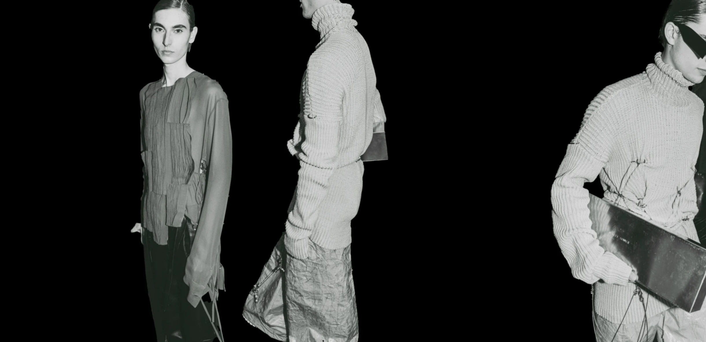 Black and white photo of three fashion mannequins dressed in modern, high-fashion clothing against a black background, with one holding a skateboard.