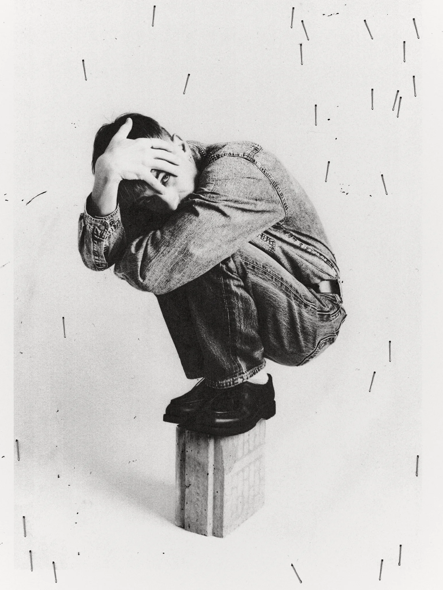 A monochrome photograph of a person crouched on a wooden block with their head resting on their hand, their face partially obscured by their hand, and one eye visible through a gap in fingers. The person is wearing a denim jacket and jeans, and is positioned in front of a plain wall with scattered objects resembling nails or pins.