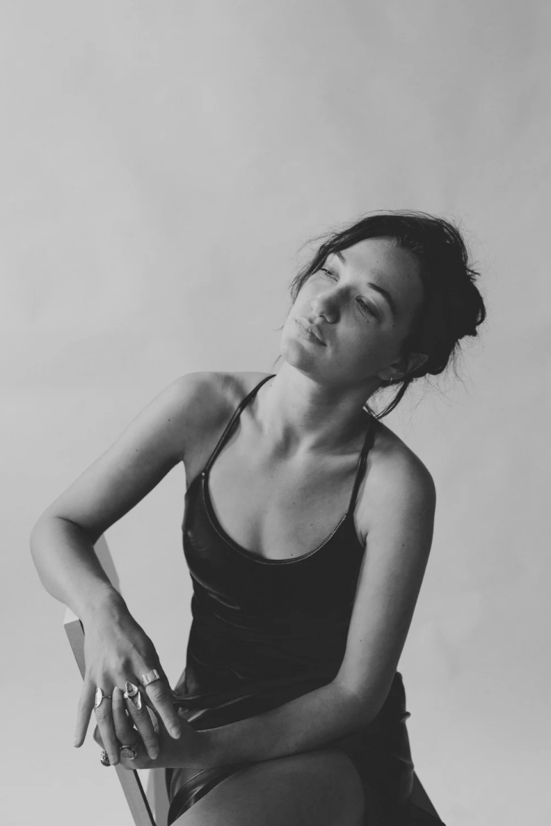Black and white photo of a woman with short, tousled hair, wearing a satin slip dress, sitting on a chair, looking off to the side with a contemplative expression, against a plain background.