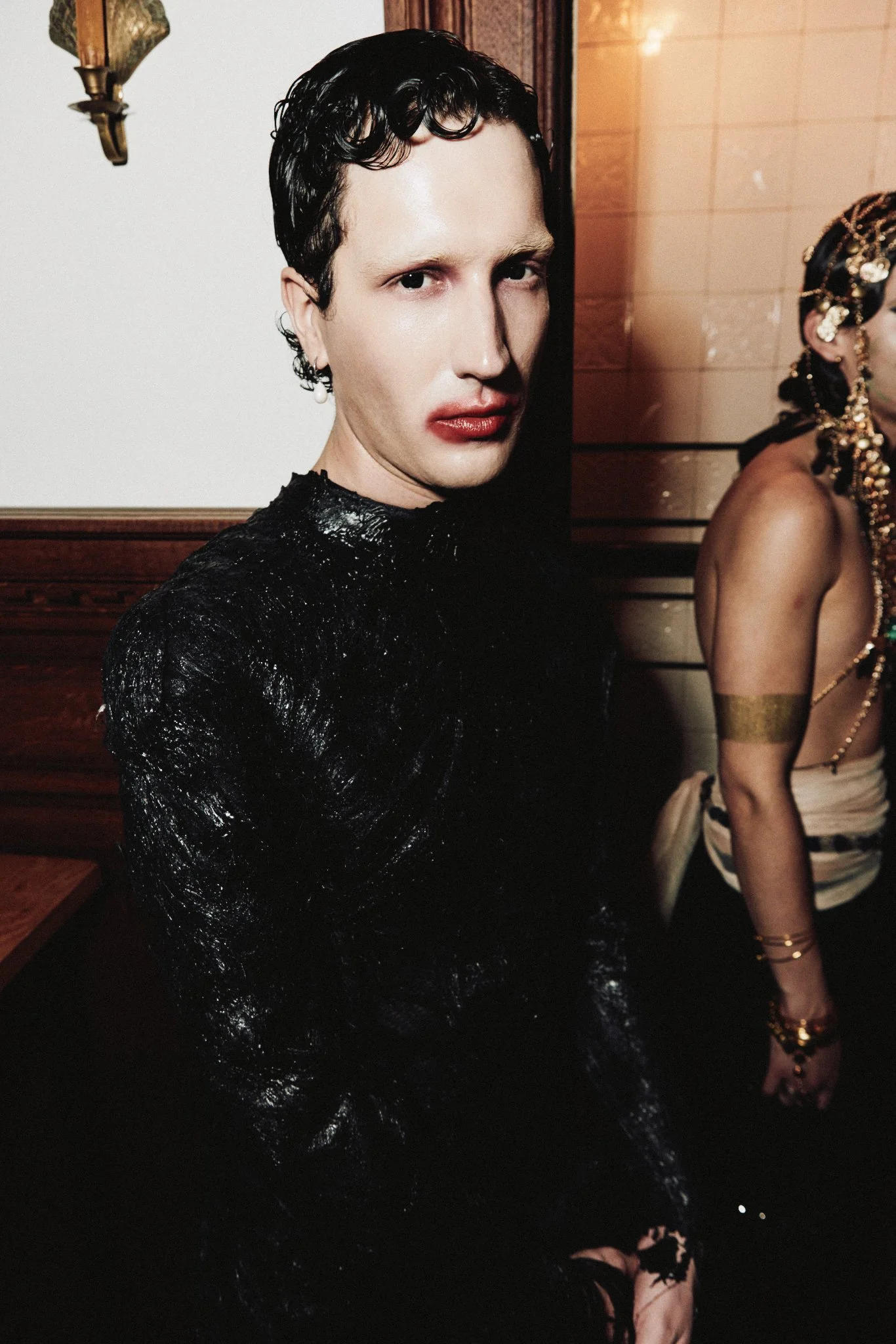 A person with pale skin and short dark curly hair, wearing makeup with red lipstick, dressed in a shiny black outfit. Partially visible person to the right wearing traditional jewelry and clothing, sitting against a wood-paneled wall.