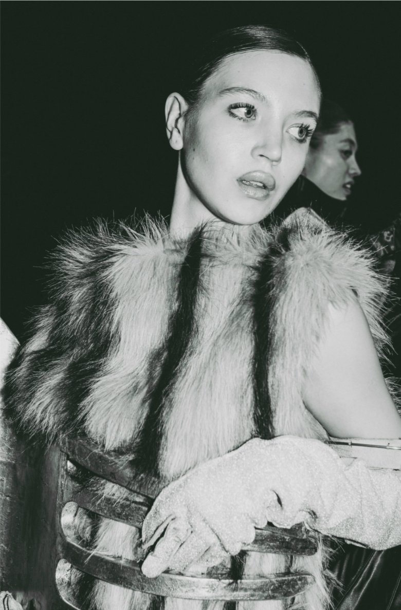 A young woman with light skin, light-colored eyes, wearing a fur stole and elegant gloves, sitting on a chair in a black and white photograph, with another woman blurred in the background.