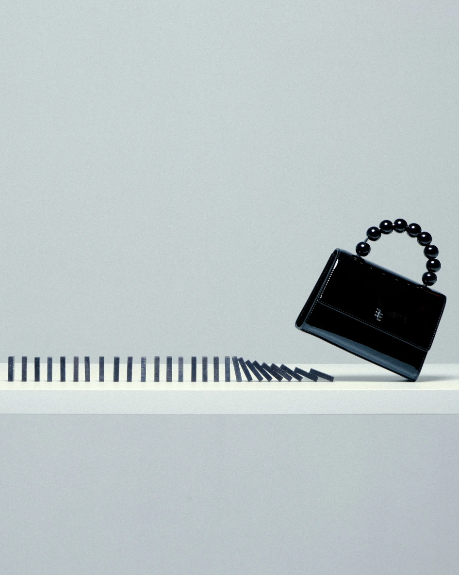 A black handbag with a bead handle, placed on a white surface next to a row of black dominoes, some of which are falling.