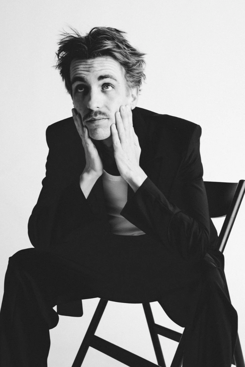 A black-and-white photo of a man sitting on a chair with a contemplative expression, hands on his cheeks, and wearing a dark blazer over a white shirt.