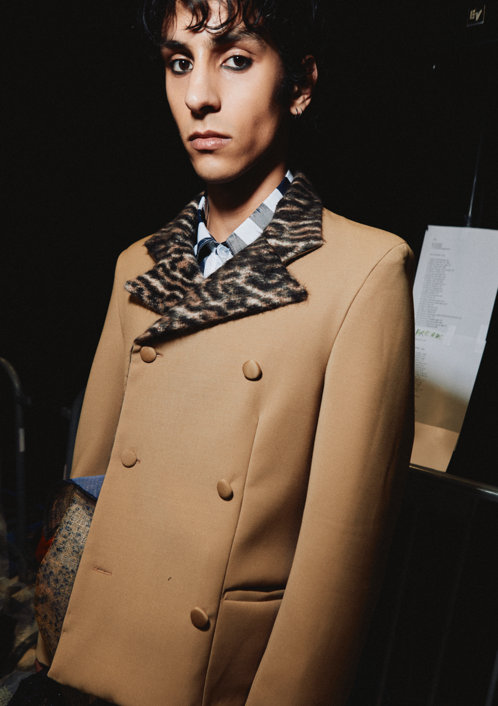 A person with short, dark, curly hair wearing a beige coat with a leopard print collar and black buttons, a patterned shirt underneath, and earrings, standing against a dark background.