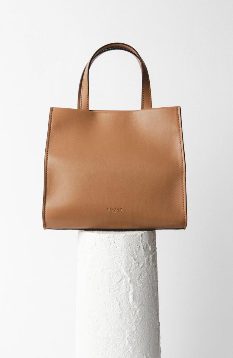 Brown leather tote bag with the brand name VOORT embossed near the bottom center, sitting on a white textured pedestal against a plain white background.