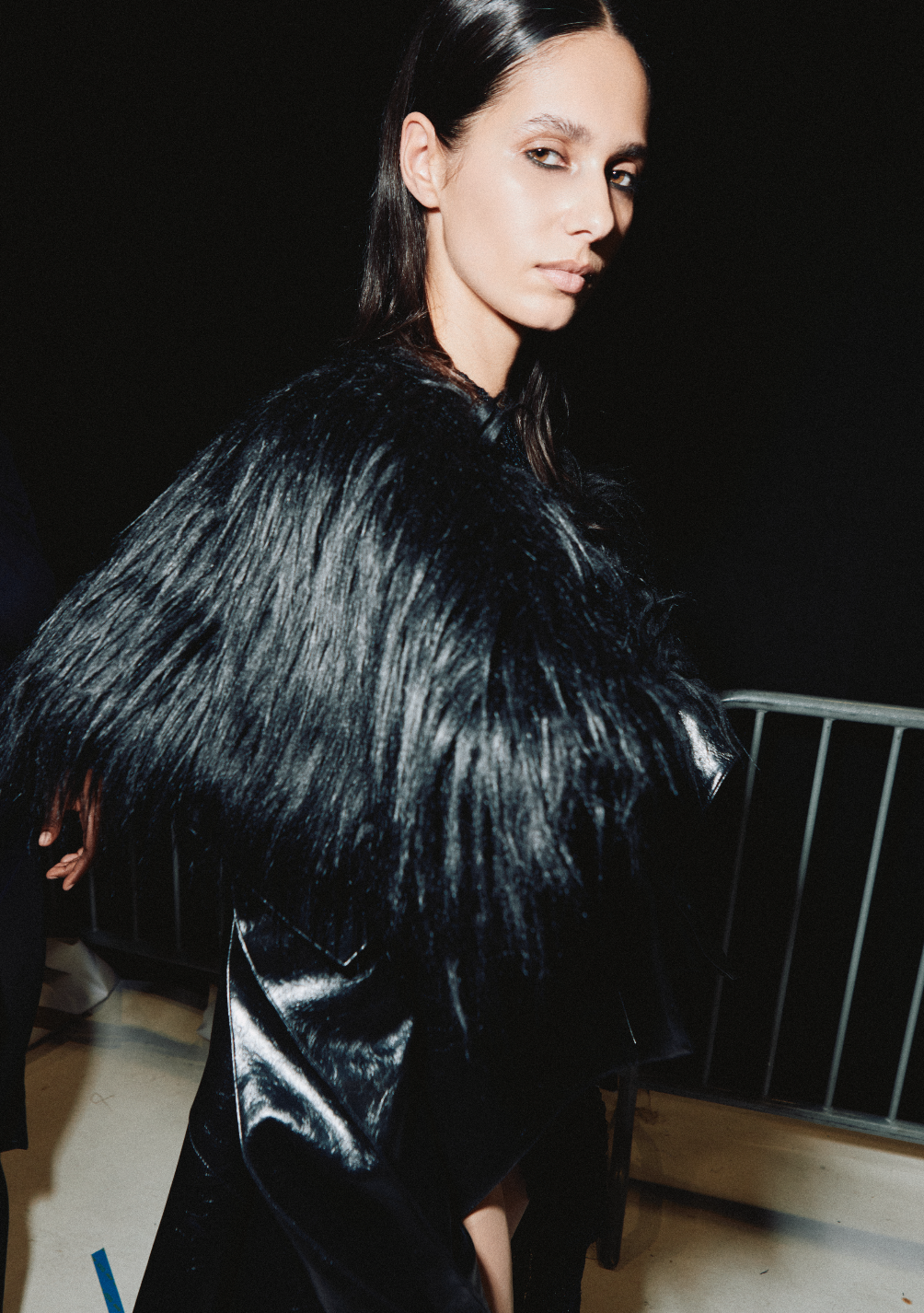 A woman with long, dark hair and a serious expression, wearing a black shiny leather jacket with a fur collar, standing in front of a dark background.