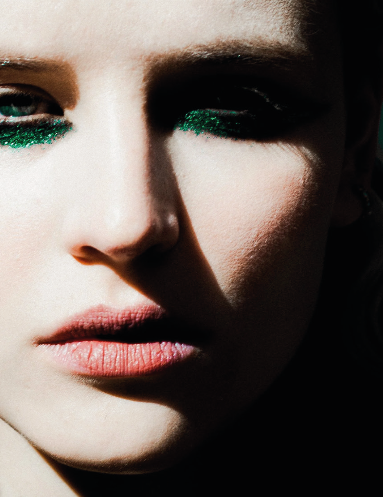 Close-up of a woman's face with makeup featuring green glitter on her eyelids, pale skin, and pink lips.
