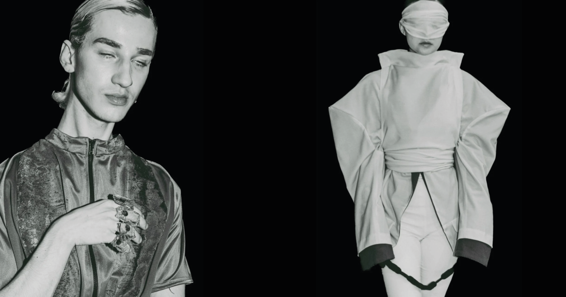 Black-and-white photo of a woman in a shiny athletic jacket holding a small object, and a person dressed in traditional Asian clothing with a blindfold covering their eyes, standing against a black background.