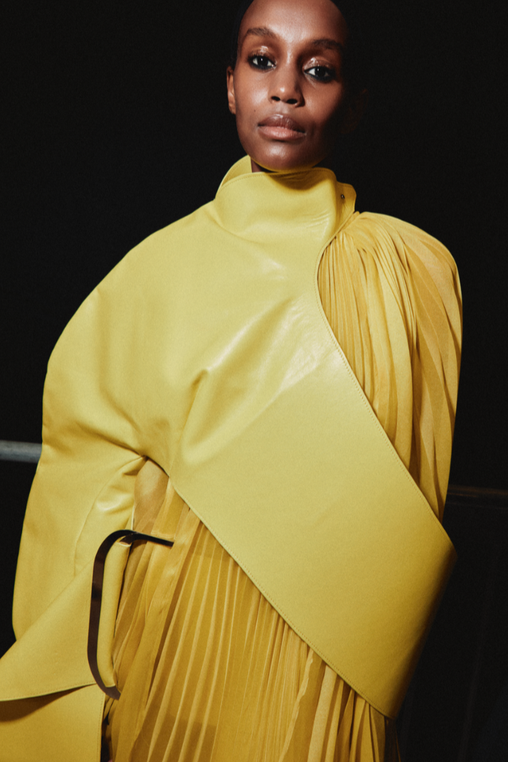 A woman with dark skin and short black hair wears a unique, yellow fashion look with a high collar, draped fabric, and pleated sections, standing against a dark background.