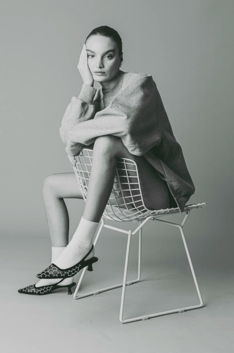 Black and white photo of a woman sitting on a wire chair with her left elbow resting on her knee, her chin resting on her hand, wearing a casual sweatshirt, patterned kitten heel shoes, and white socks.