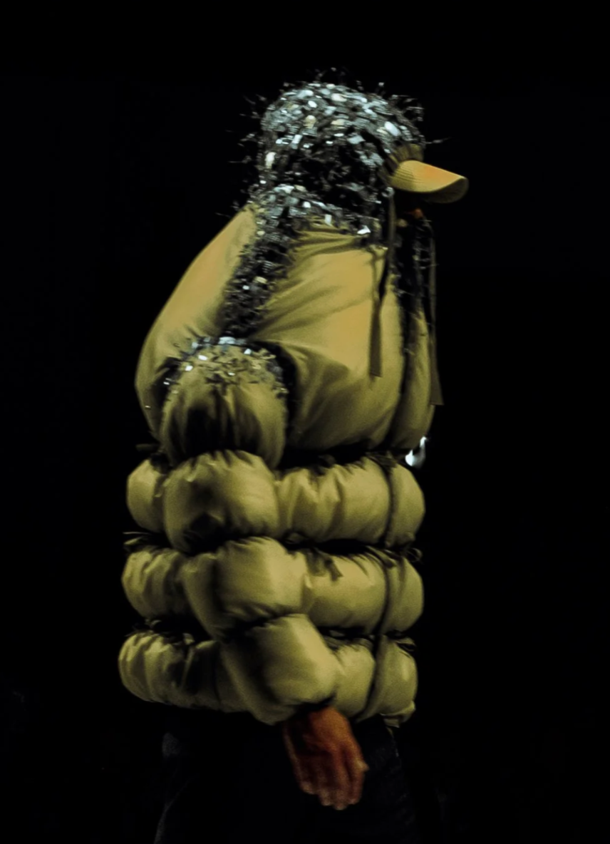 Person in a yellow puffy jacket with reflective embellishments, wearing a yellow hat, standing against a dark background.